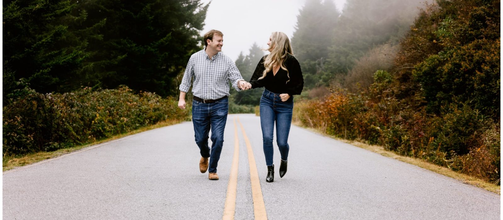 young engaged couple holding hands smiling at one another while running down the middle of a forested road for their outdoor winter engagement photo session