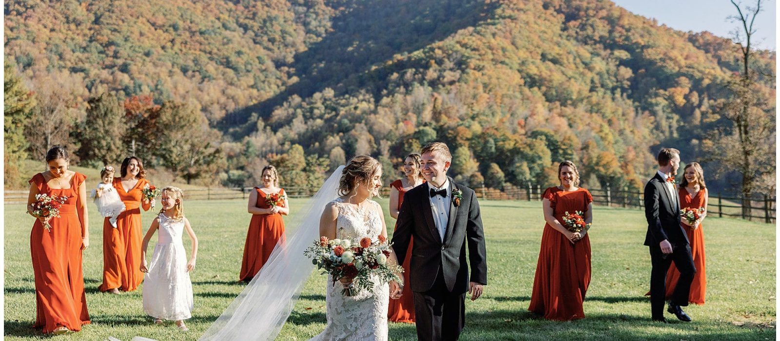 A bride and groom walk hand-in-hand at their outdoor wedding, with bridesmaids in orange dresses and groomsmen in the background, all against a backdrop of autumn mountains.