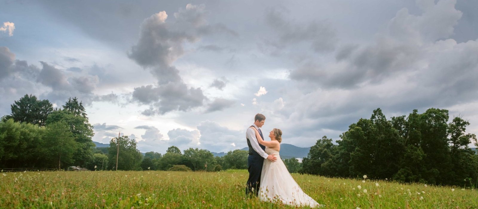 Groom standing in front of bride holding her hips while standing in grassy field smiling at one another